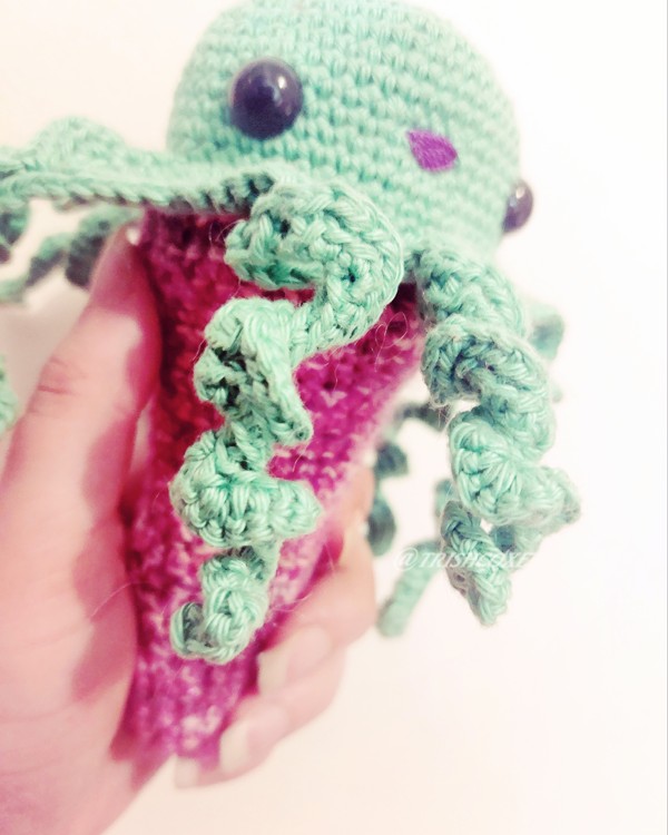An octopus crocheted with green yarn, with black round eyes, a purple embroidered mouth, and curly dangling arms hanging over a magenta crochet 'cone'. 